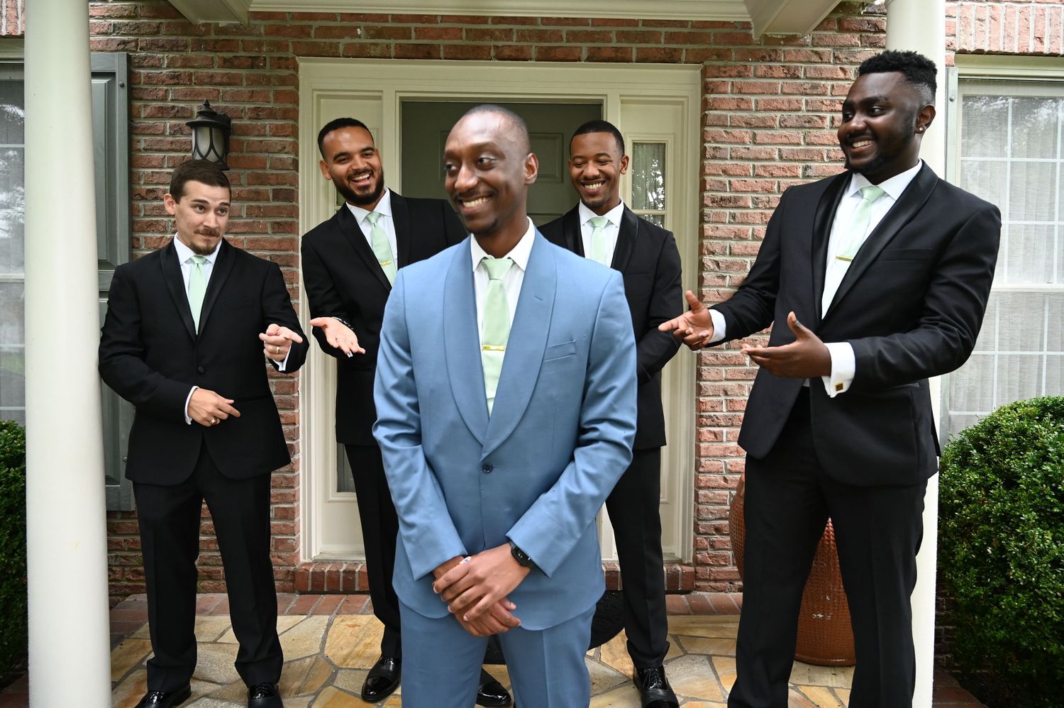 Groom in light blue suit laughing with four groomsmen in black tuxedos outside a brick home.