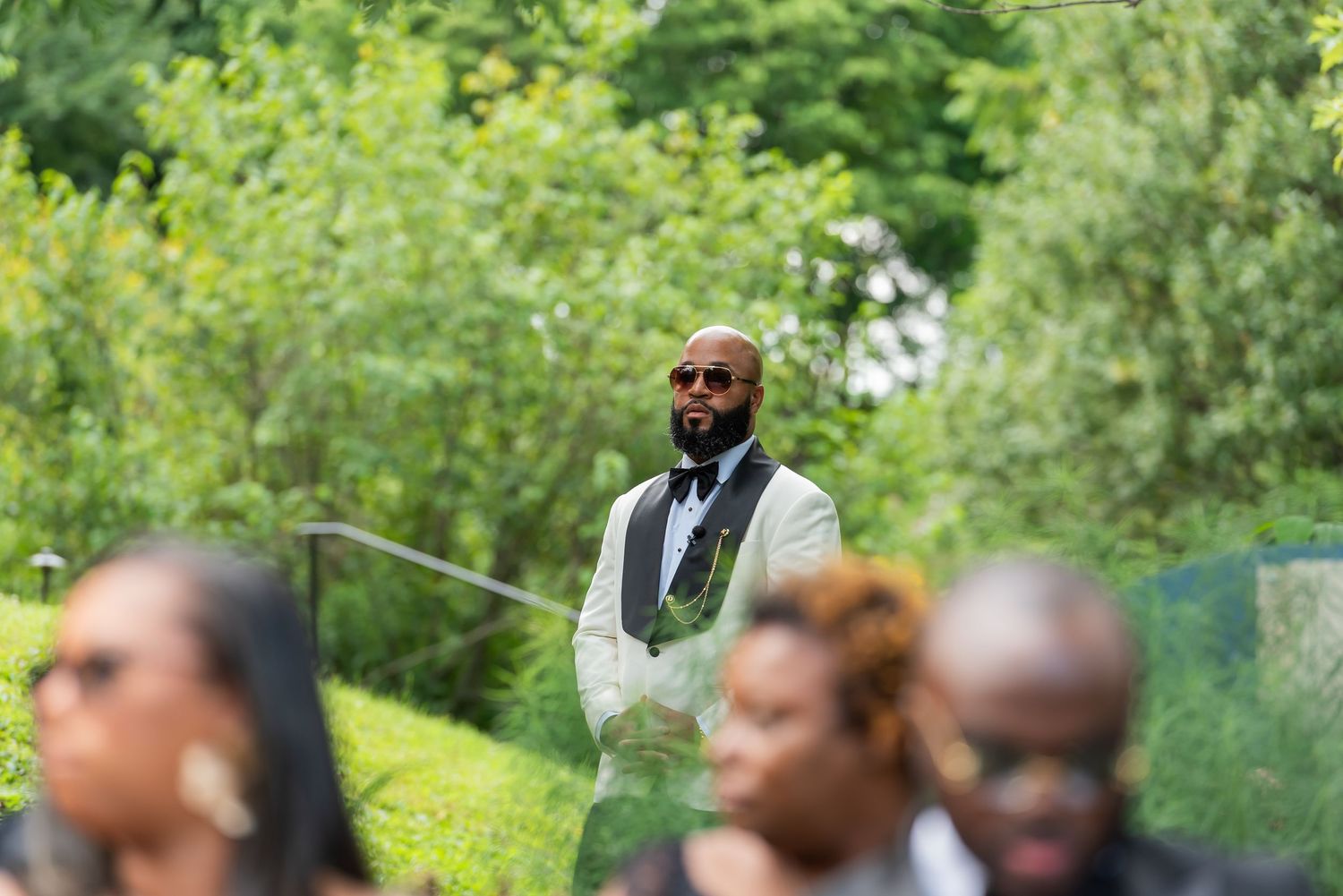 A well-dressed bald man in a vest and sunglasses stands outdoors at a wedding ceremony surrounded by lush green trees.