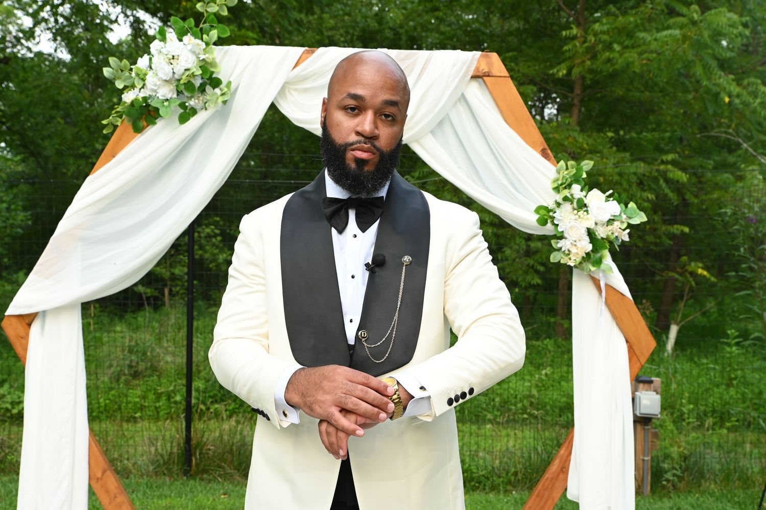 Elegant man in white tuxedo with black lapels stands before floral arch at outdoor wedding ceremony.