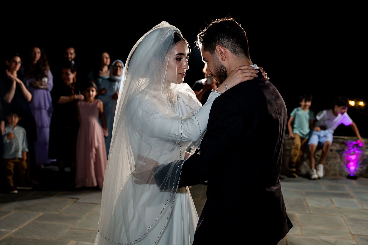 Bride and groom share first dance at outdoor night wedding ceremony with guests watching in background.
