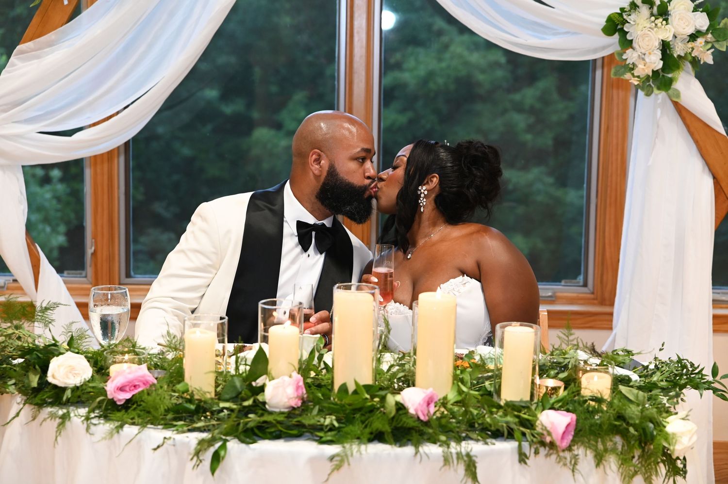 Bride and groom share a kiss at their wedding reception table adorned with candles, greenery, and pink roses.
