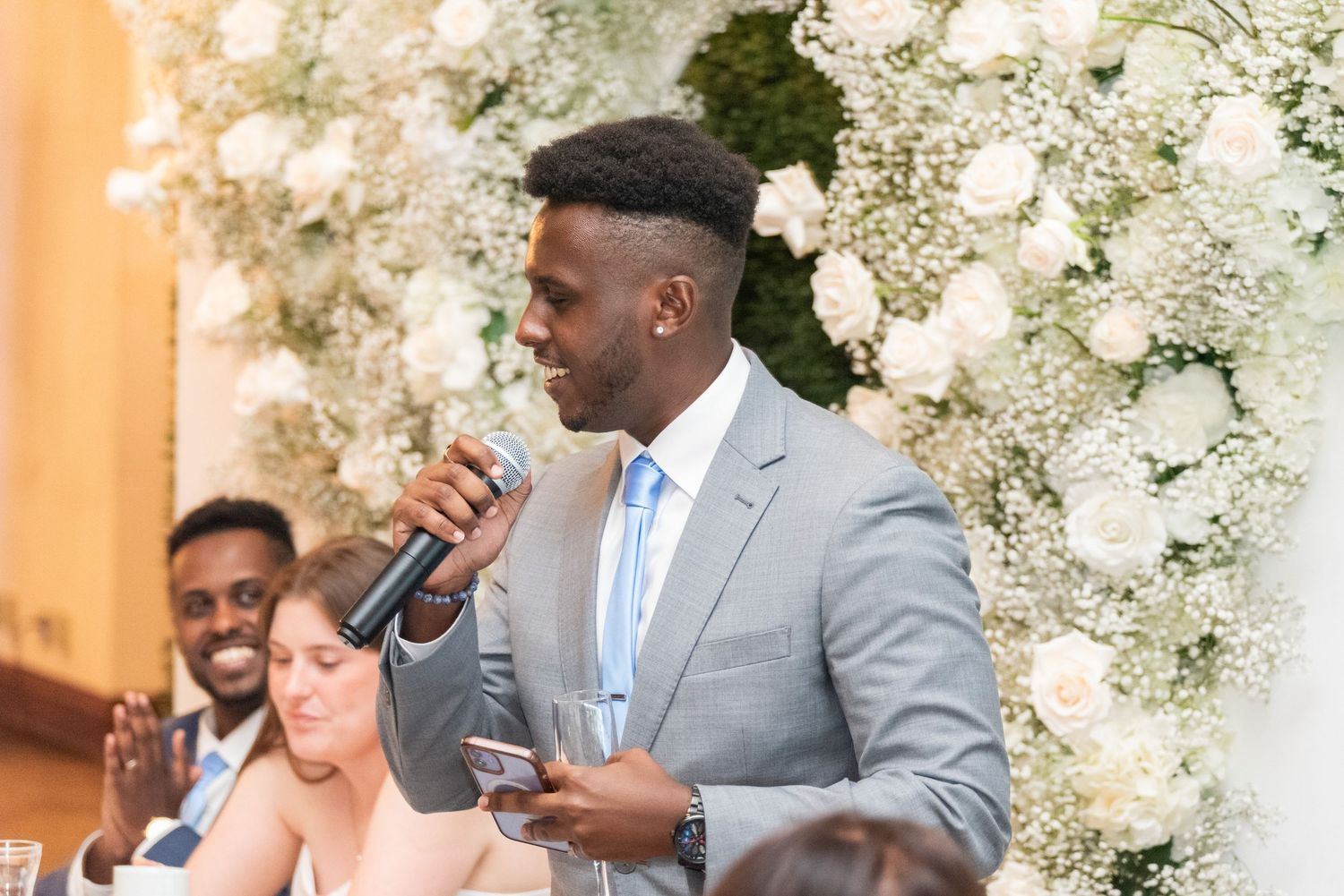 Man in gray suit giving a speech at a wedding reception with white floral arrangements in background.