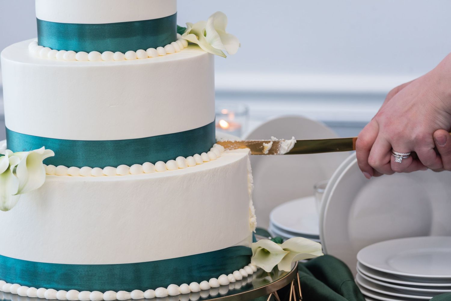 Three-tier white wedding cake with teal ribbon and calla lily decorations being cut at reception.