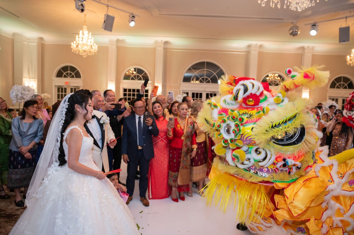 Bride in white gown watches a colorful lion dance performance at an elegant wedding reception.
