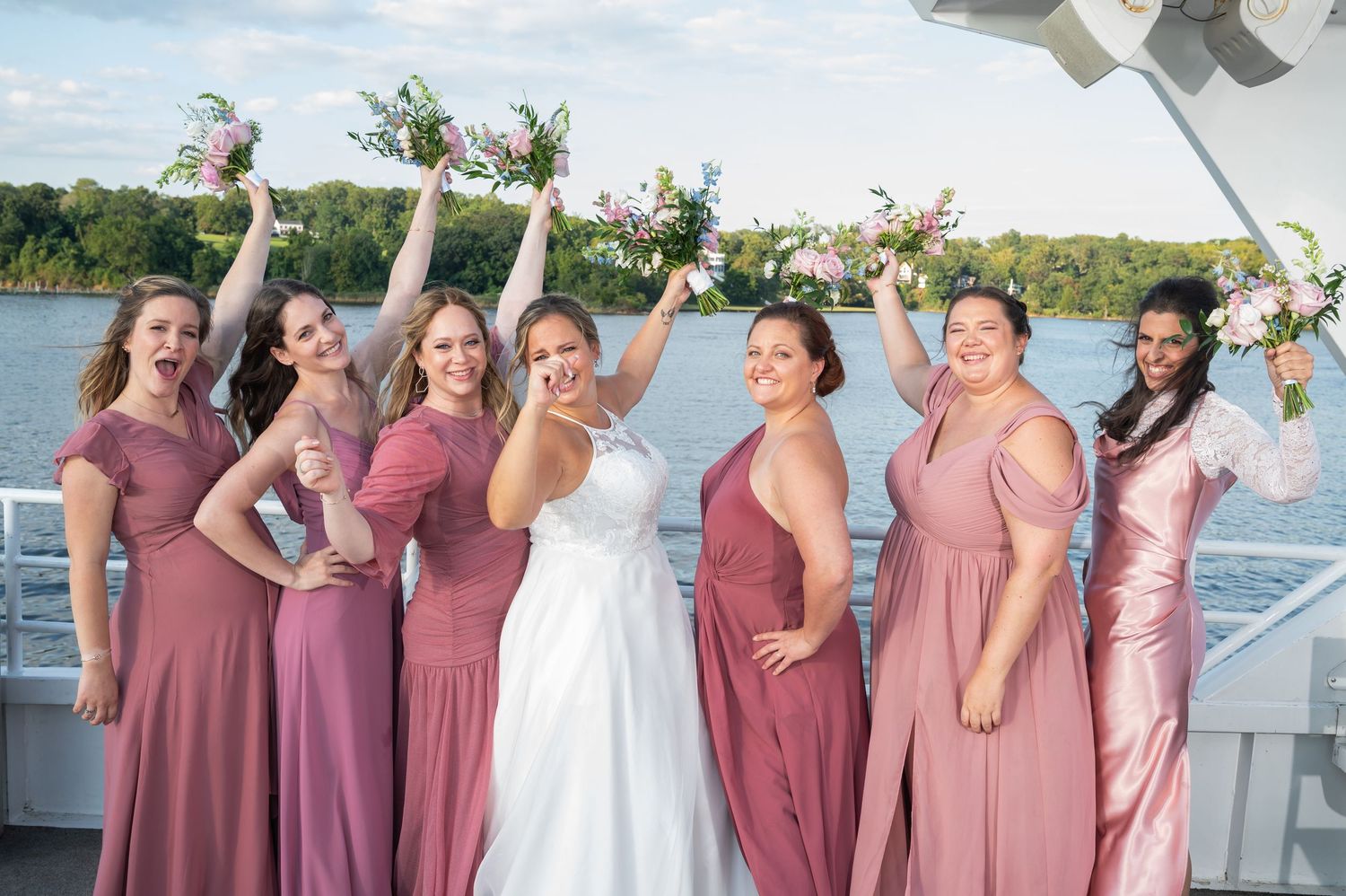 Bride with six bridesmaids in dusty rose dresses celebrating on boat deck with raised bouquets.