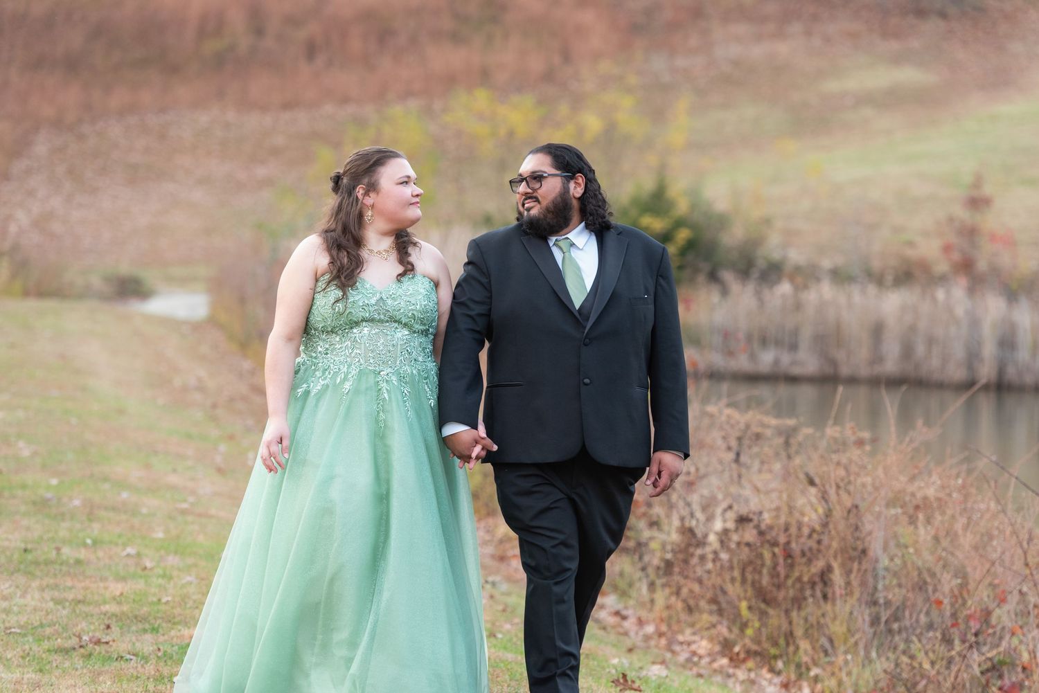 Couple in formal attire, woman in green gown and man in black suit, walking hand in hand outdoors near a pond.