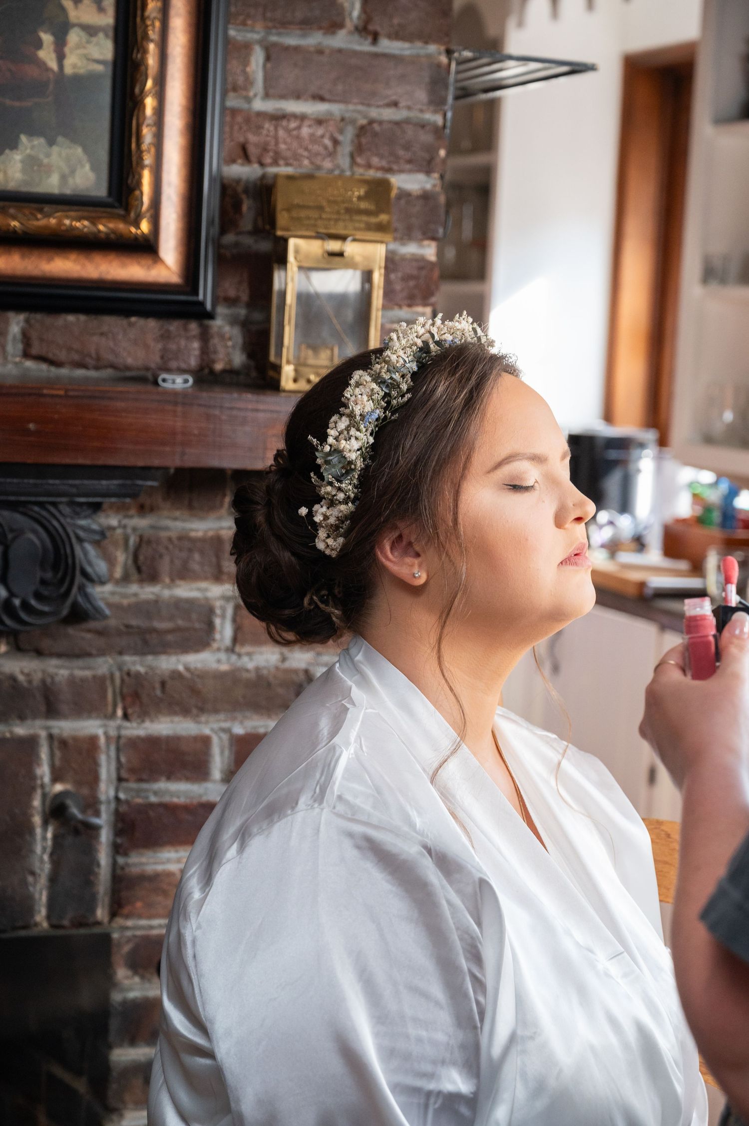 Bride in white robe with floral hair updo getting makeup applied near brick fireplace.