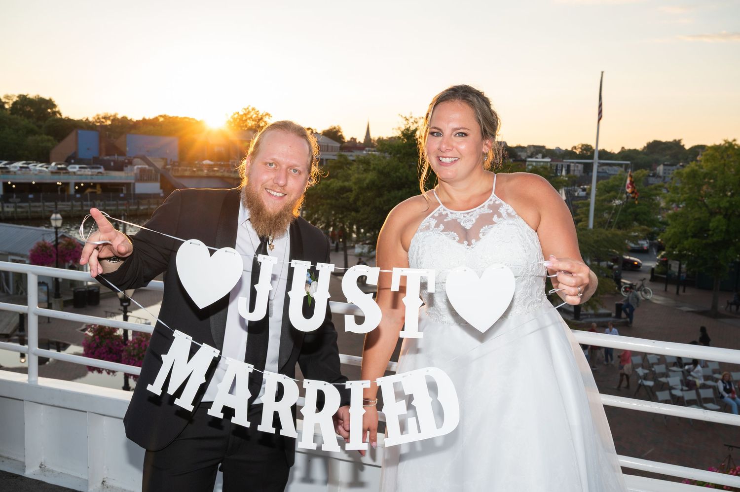 Newlyweds holding Just Married sign on bridge at sunset, bride in halter gown, groom in casual attire.