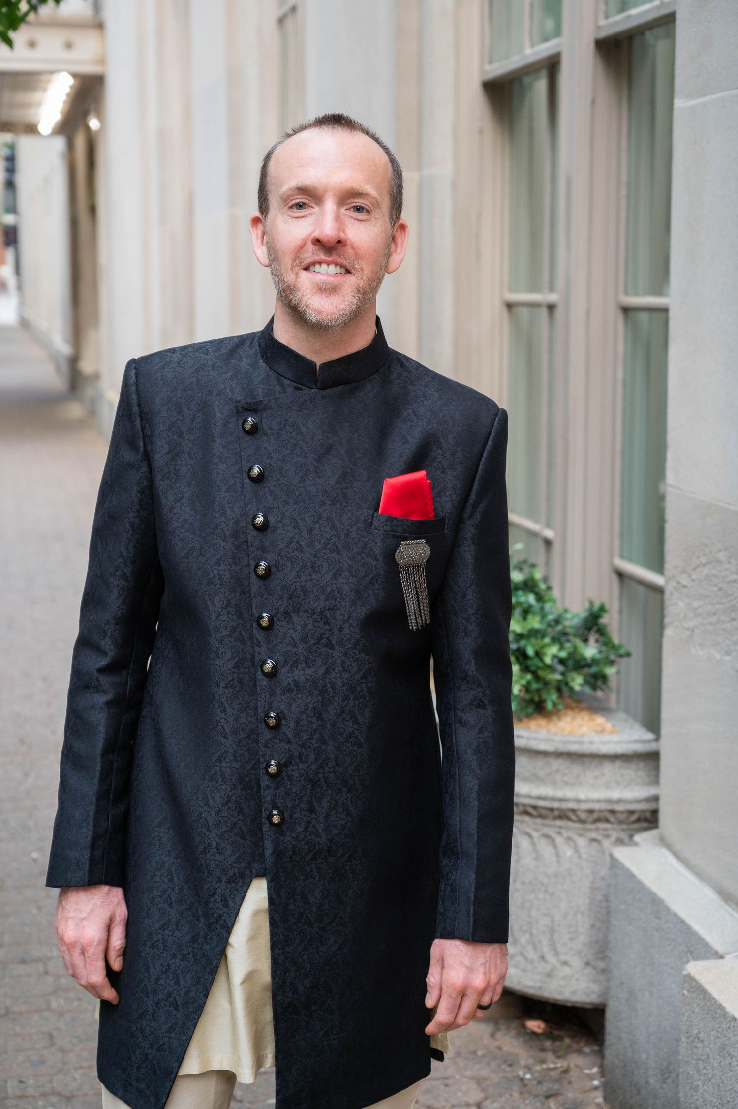 Groom in black embroidered sherwani with red pocket square posing outdoors before wedding.