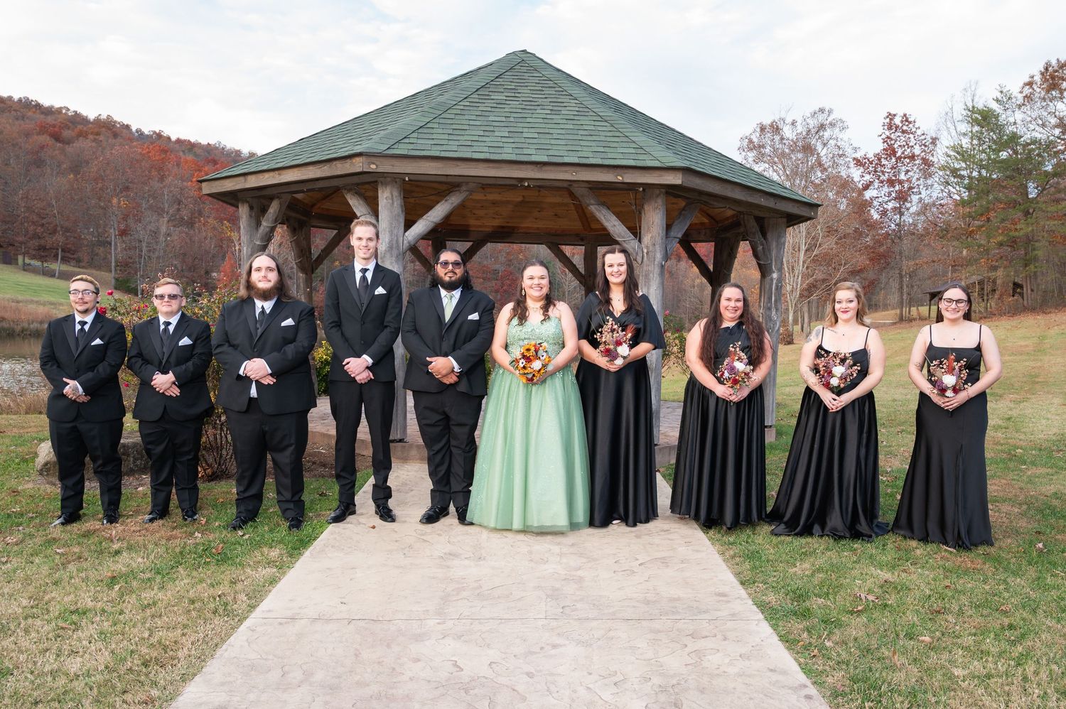 Wedding party poses before a gazebo in autumn, groomsmen in black suits and bridesmaids in green and black gowns.