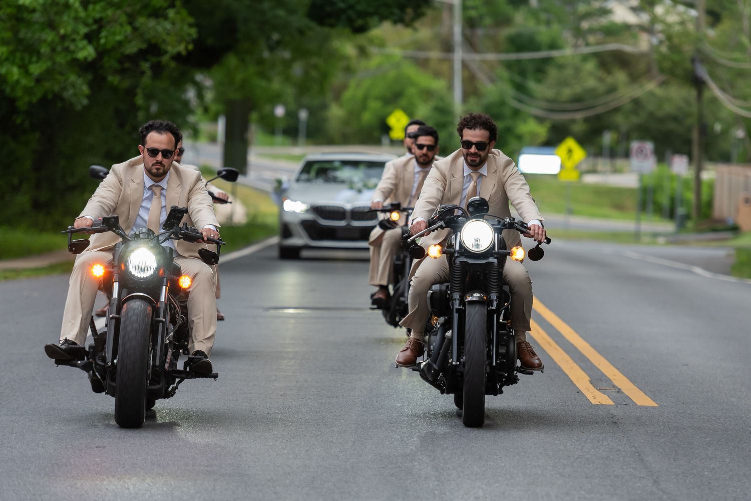 Three men in tan suits ride motorcycles side by side down a suburban road with a car following behind.