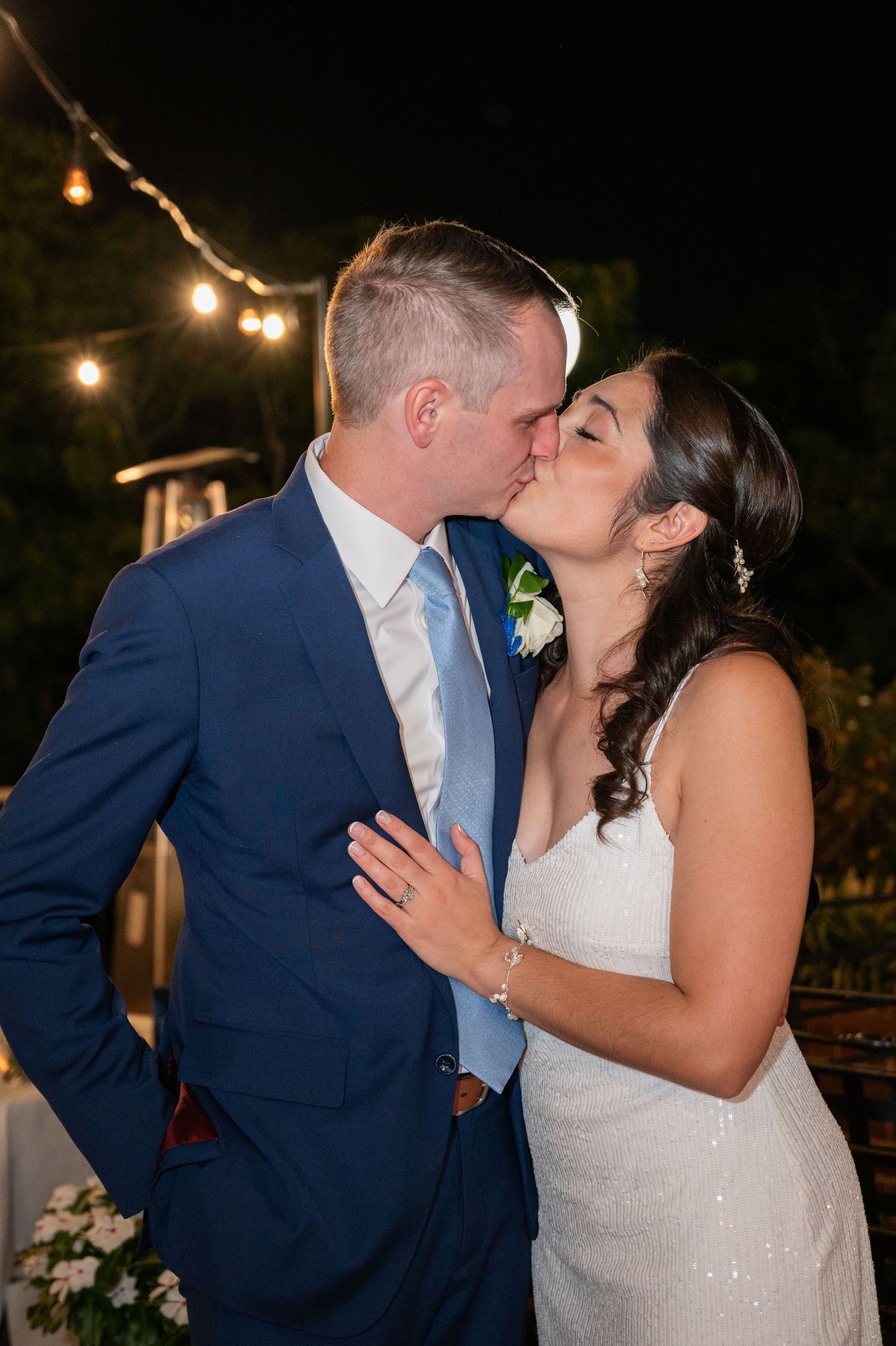 Bride and groom sharing a romantic kiss outdoors at night under warm string lights.