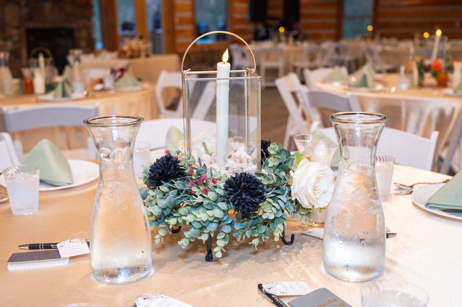 Elegant wedding reception centerpiece with candle lantern, white roses, and blue pine cones.