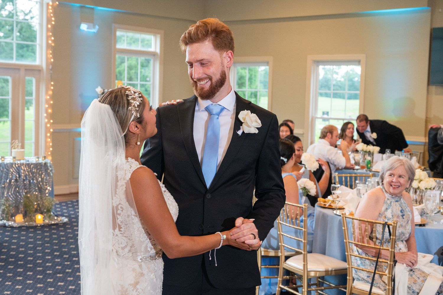 Bride and groom share first dance at elegant wedding reception with blue decor and guests seated nearby.