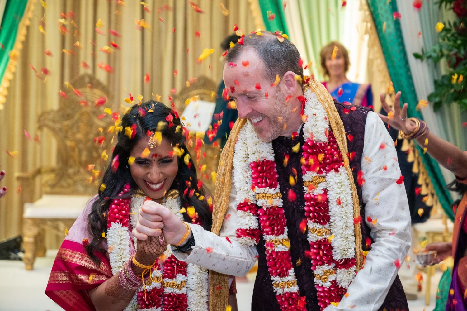 Couple celebrates Indian wedding ceremony with colorful flower petals and traditional floral garlands.