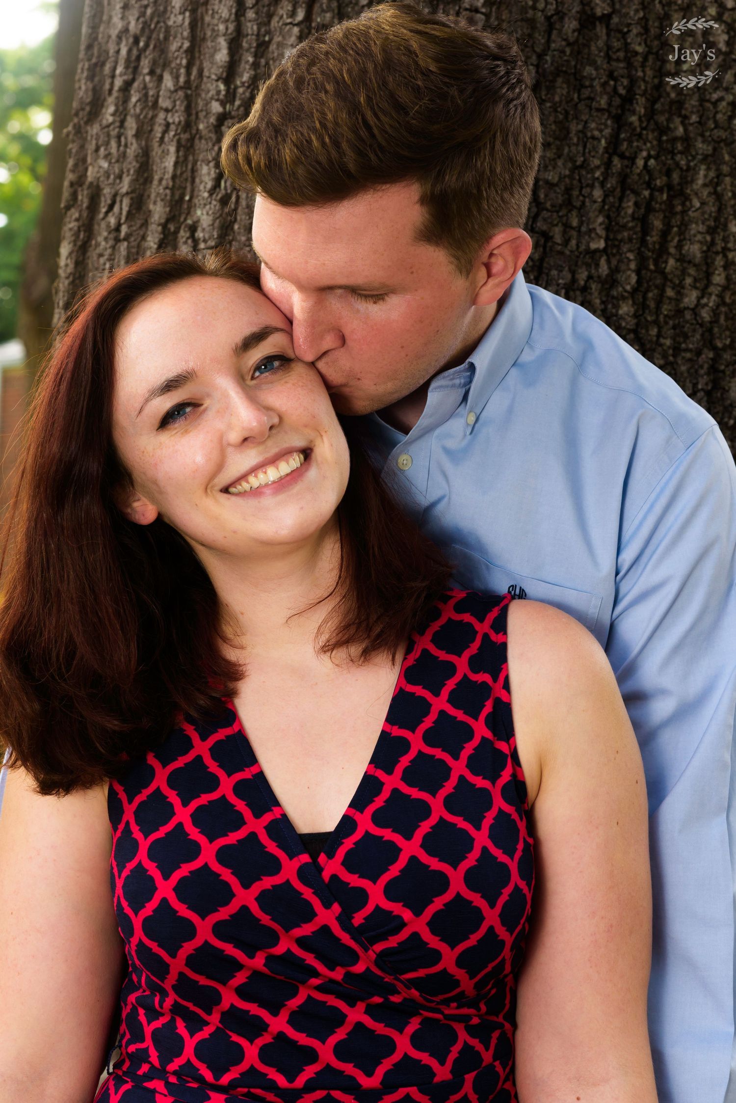 Man kissing smiling woman on forehead near a large tree outdoors.