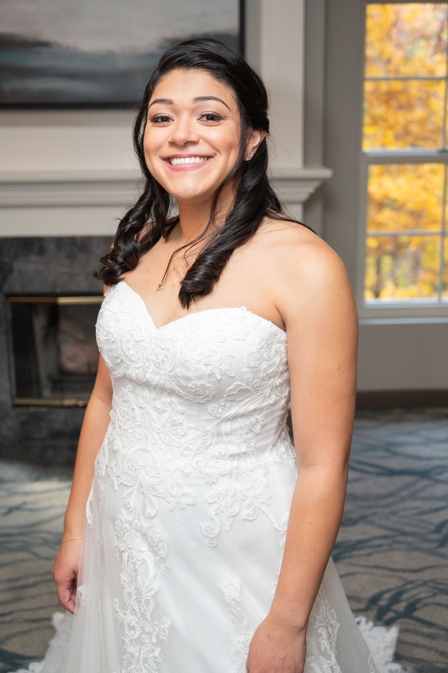 Smiling bride in strapless lace wedding dress stands near fireplace in autumn wedding venue.