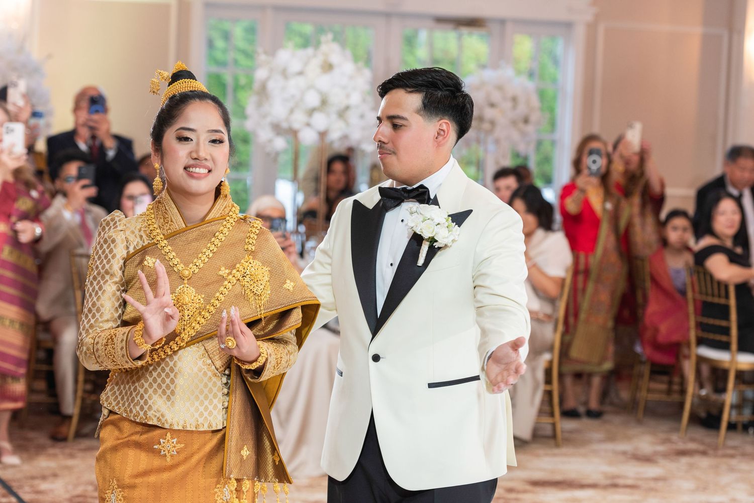 Bride in traditional golden Thai dress and groom in white tuxedo make grand entrance at elegant wedding reception.