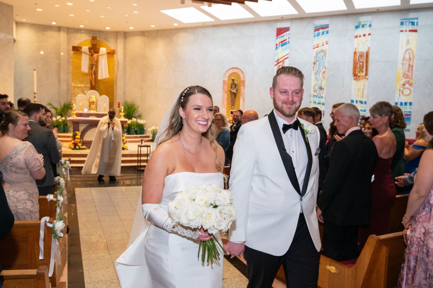 Joyful newlyweds walking down church aisle after ceremony, bride holding white bouquet.