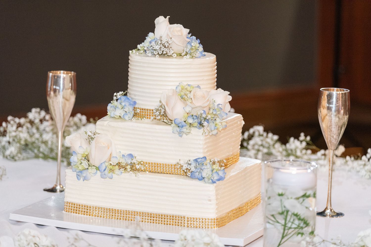 Three-tier square wedding cake with white frosting, gold ribbon trim, and blue and white floral decorations beside champagne flutes.