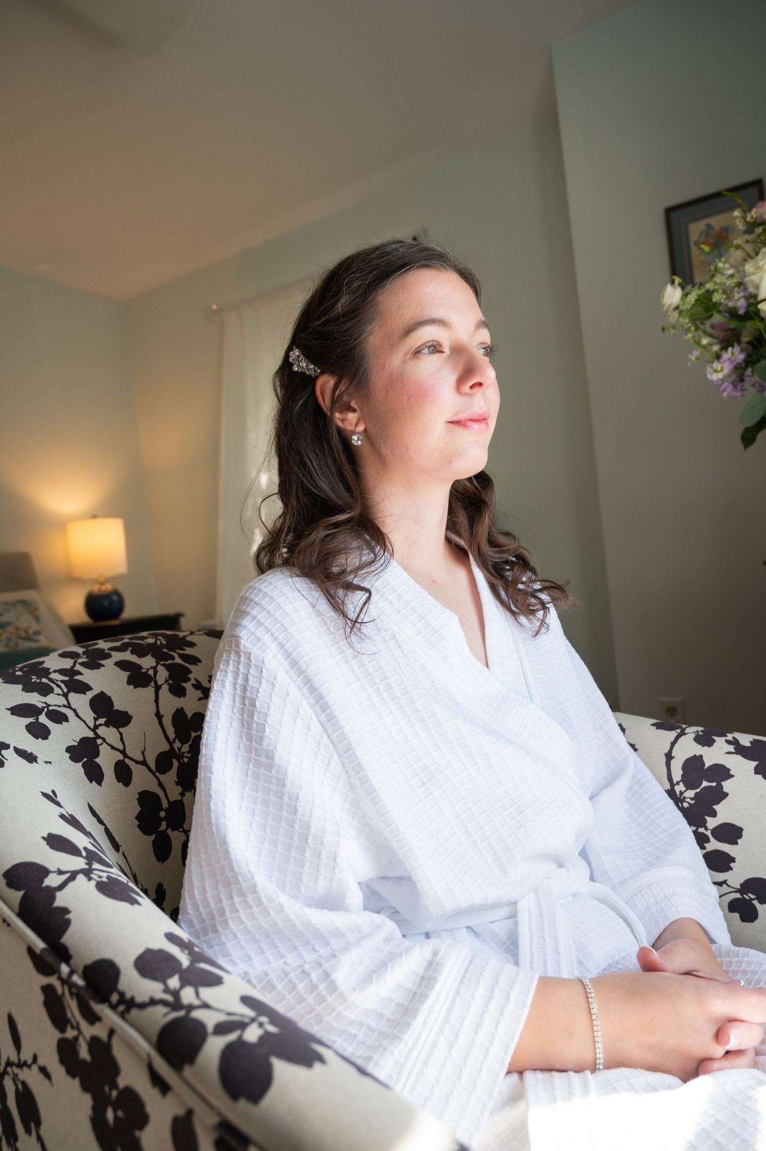 Bride in white robe sitting on floral chair gazing thoughtfully on her wedding morning.