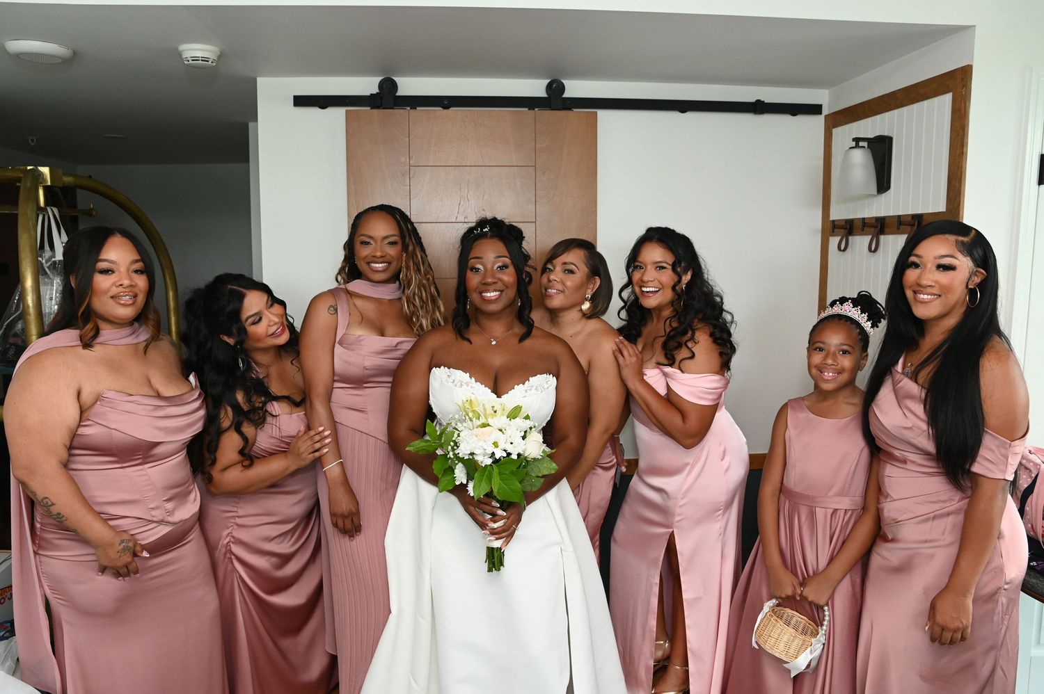 Bride in white gown holds bouquet surrounded by six bridesmaids in dusty rose satin dresses, smiling indoors.