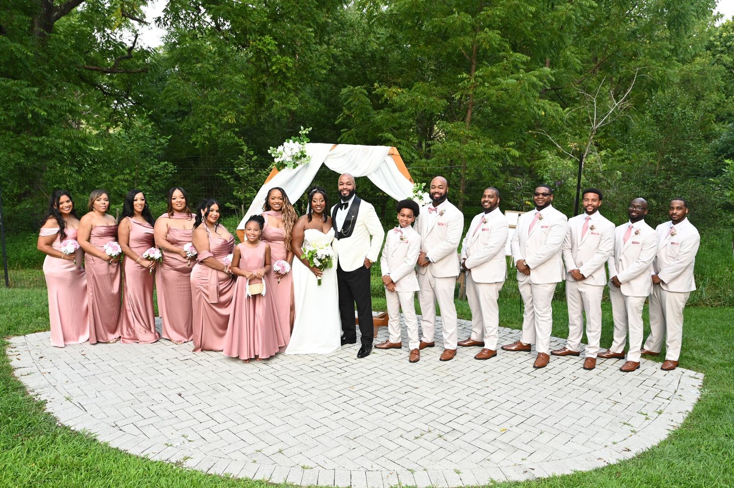 Wedding party poses outdoors with bridesmaids in pink dresses and groomsmen in white suits on a brick patio.