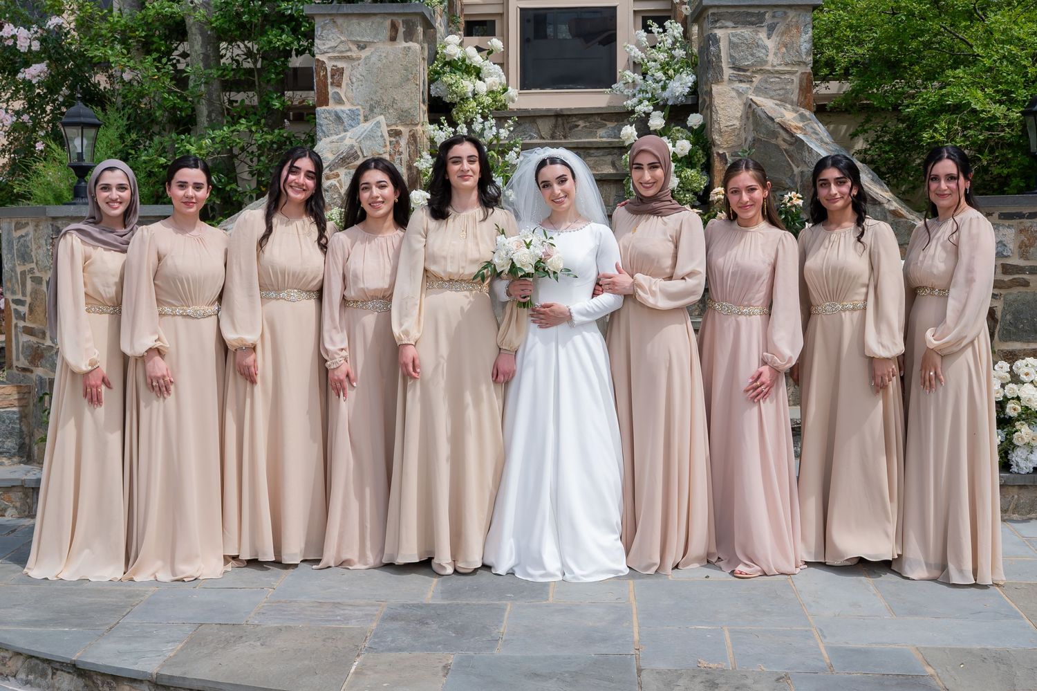 Bride in white gown poses with bridesmaids in matching champagne dresses at an outdoor wedding ceremony.