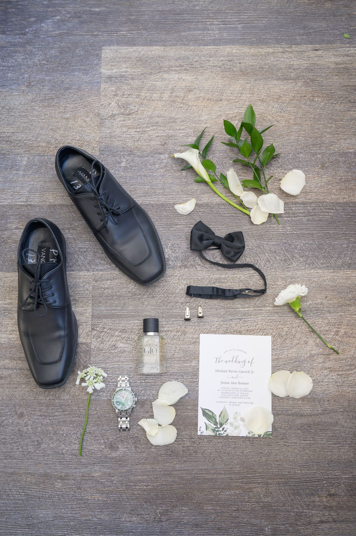 Groom wedding flat lay featuring black dress shoes, bow tie, watch, cologne, earrings, white flowers, and wedding invitation on wood floor.