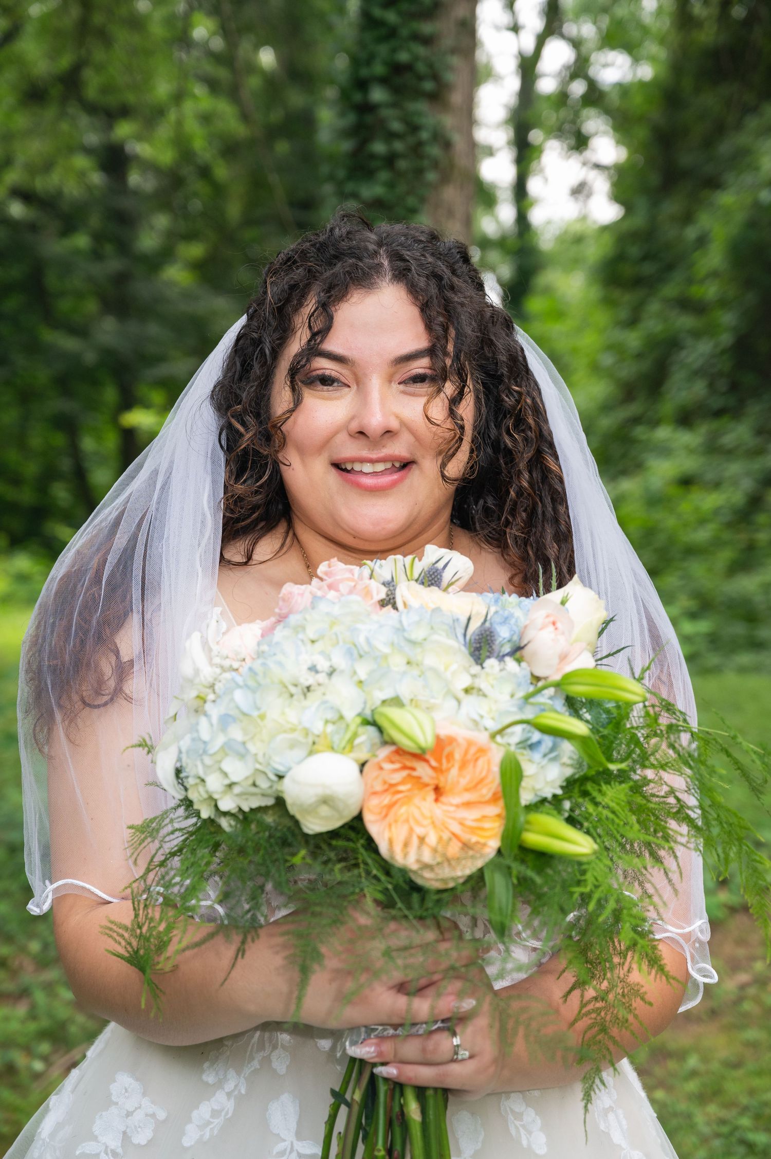 Smiling bride in lace wedding dress holds pastel bouquet with hydrangeas outdoors in garden.