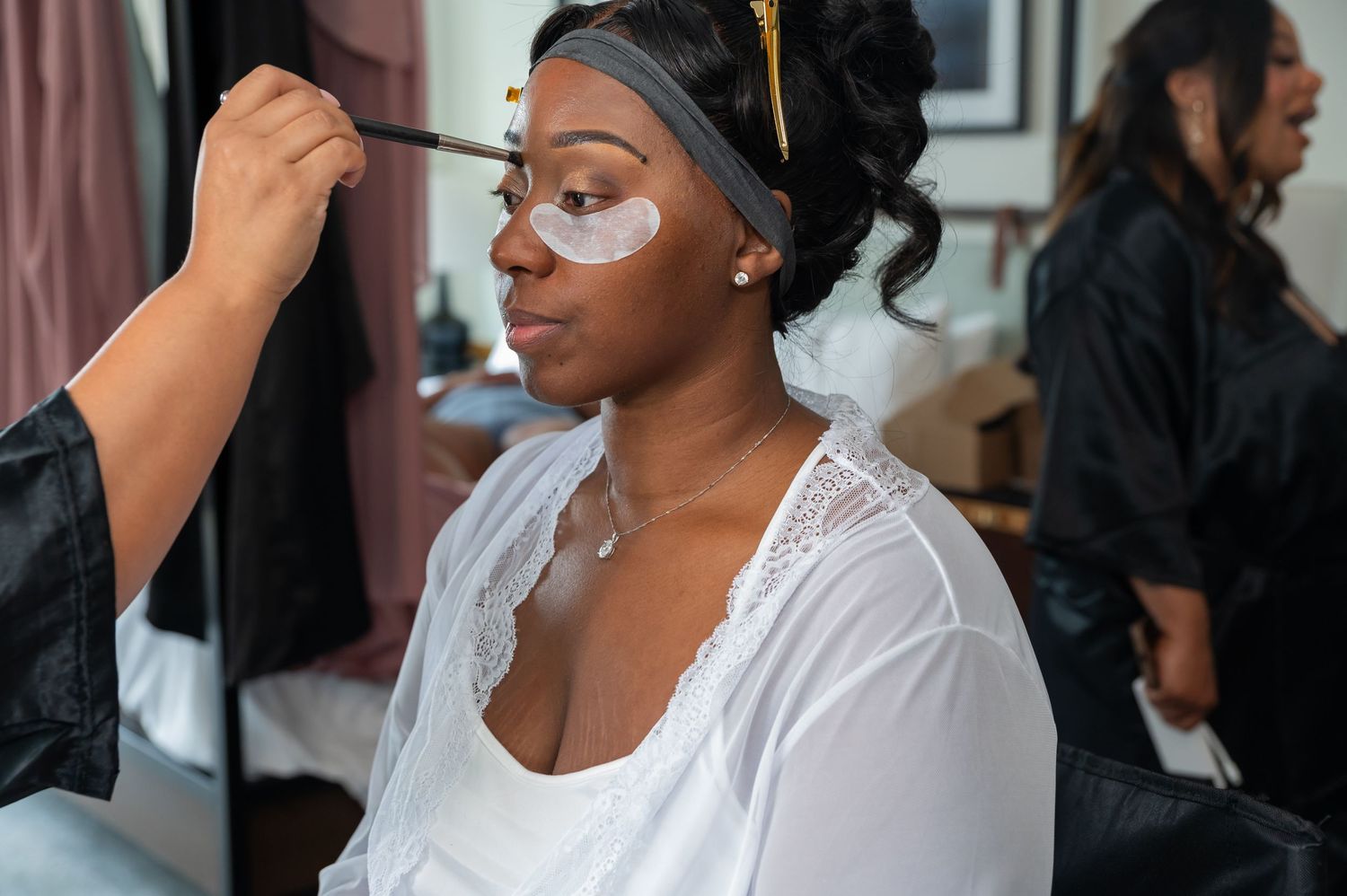 A makeup artist applies eye makeup to a bride wearing under-eye patches and a white robe during wedding preparations.