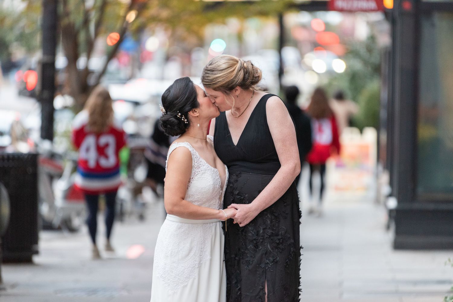 Two brides sharing a tender moment on a city sidewalk outdoors.