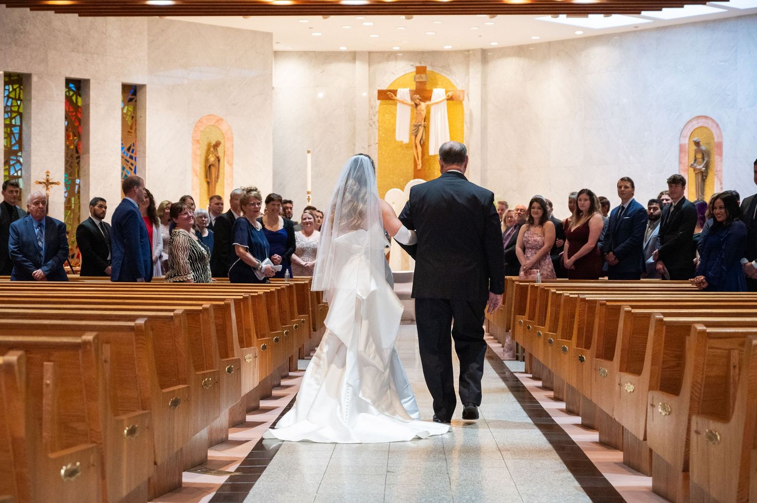 Father walks bride down church aisle in white gown and veil before seated wedding guests.