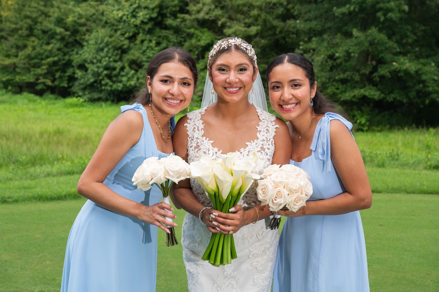 Smiling bride in lace wedding dress poses with two bridesmaids in light blue gowns holding white bouquets outdoors.