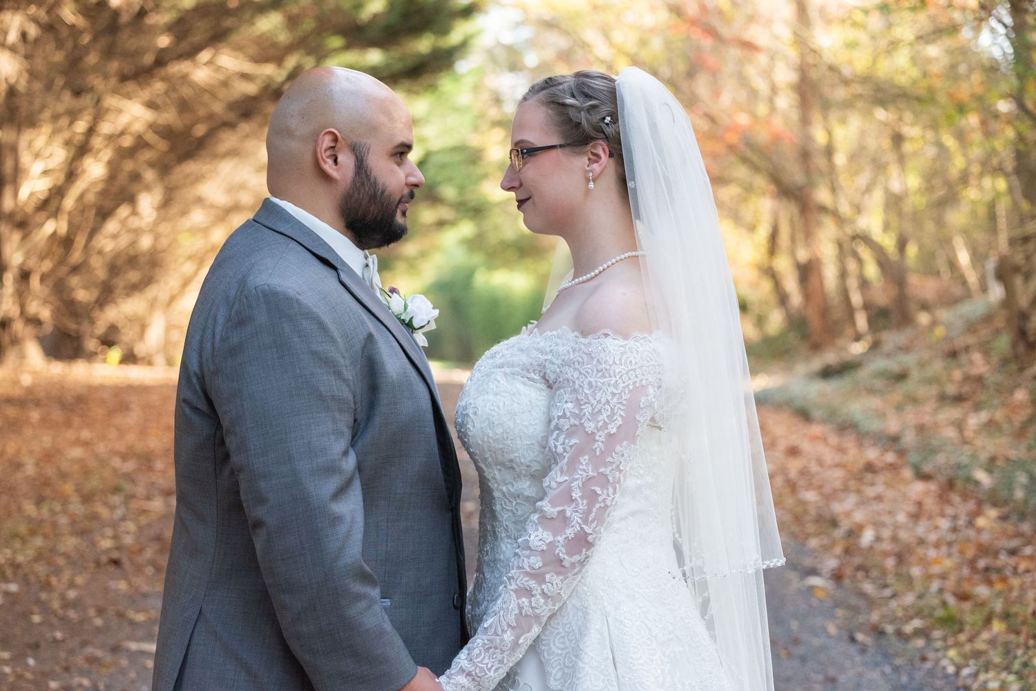 Bride and groom hold hands and gaze at each other on a tree-lined autumn path.