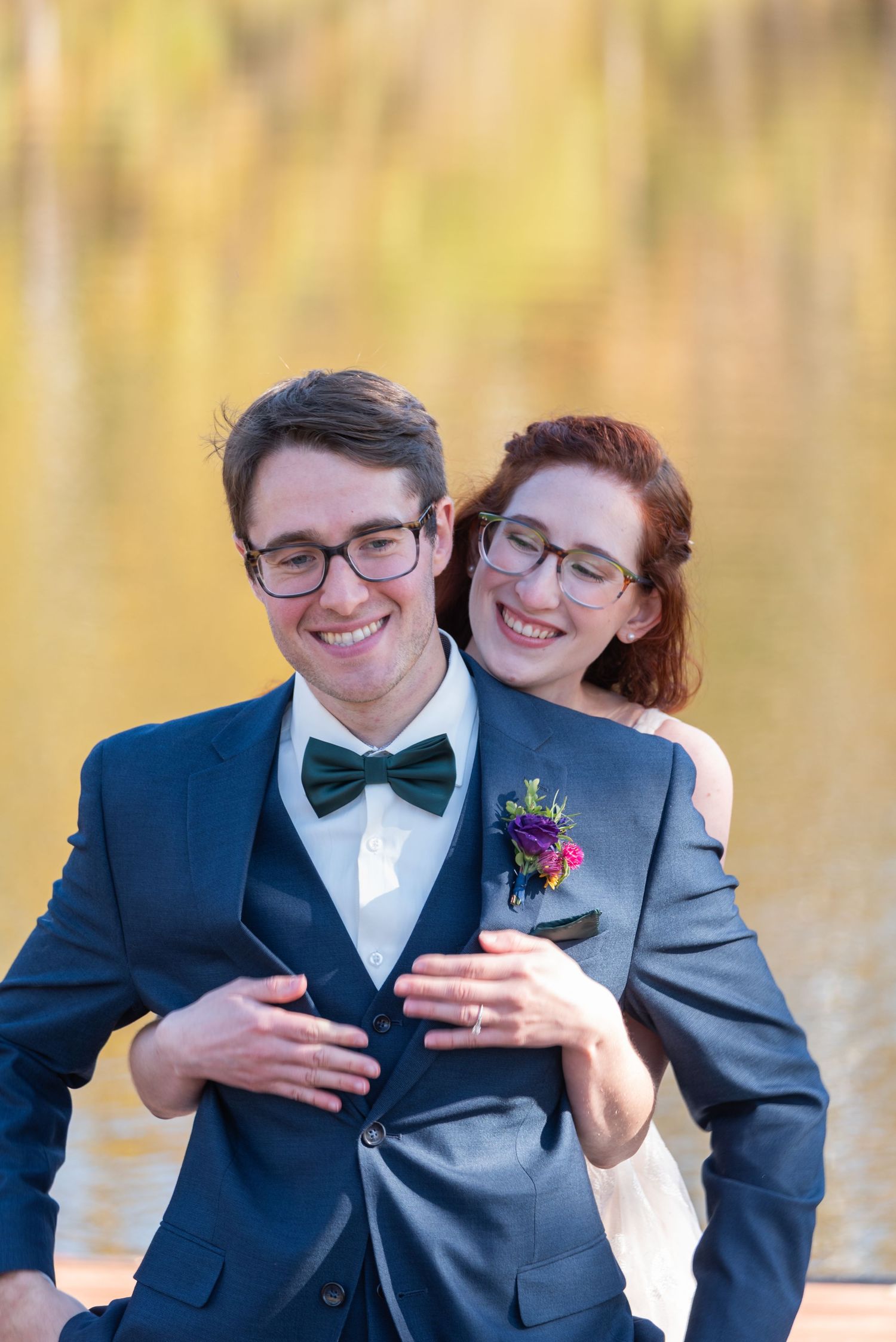 Happy couple embracing by autumn lake, groom in navy suit with bow tie and floral boutonniere, bride in gray blazer.