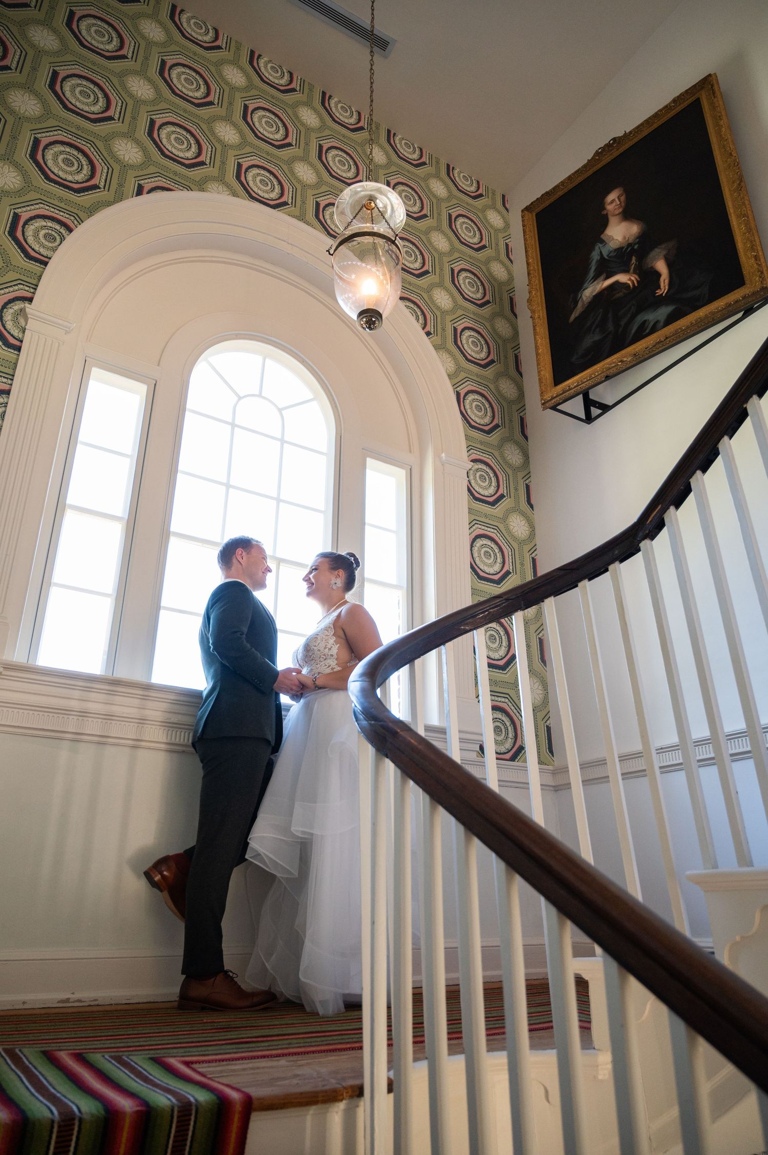 Bride and groom share a tender moment on an elegant staircase with arched window and classic portrait in a historic venue.