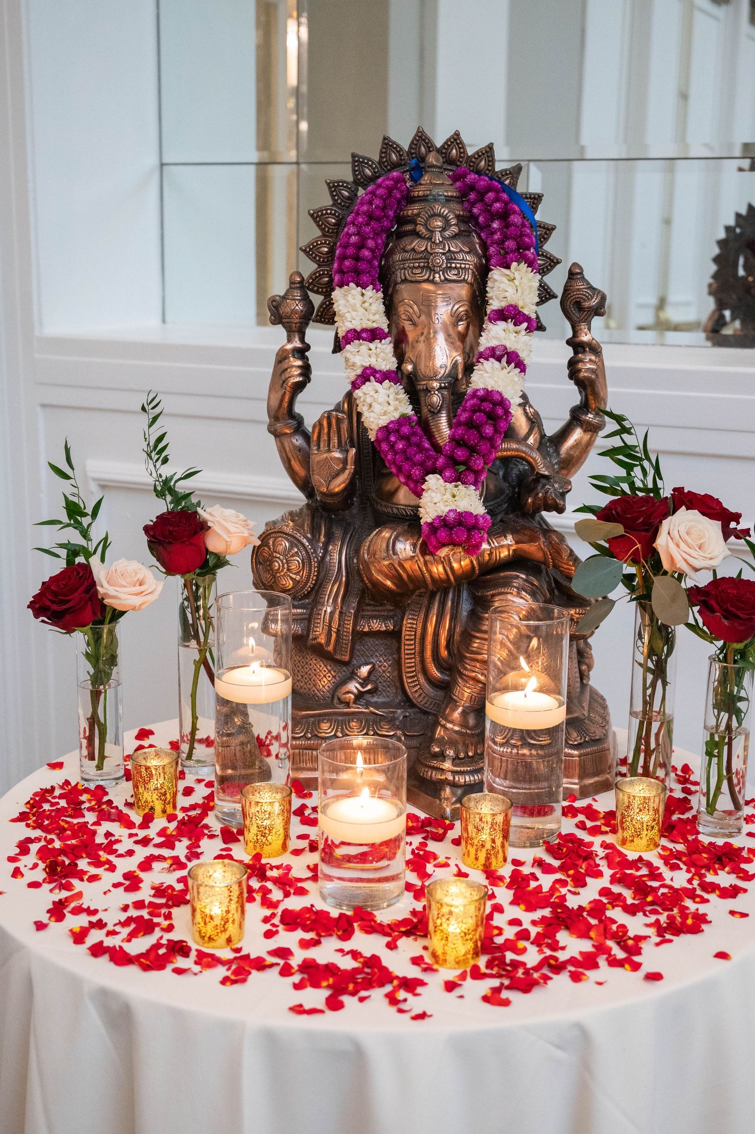 Bronze Ganesha statue adorned with floral garlands surrounded by lit candles and red rose petals on a white table.