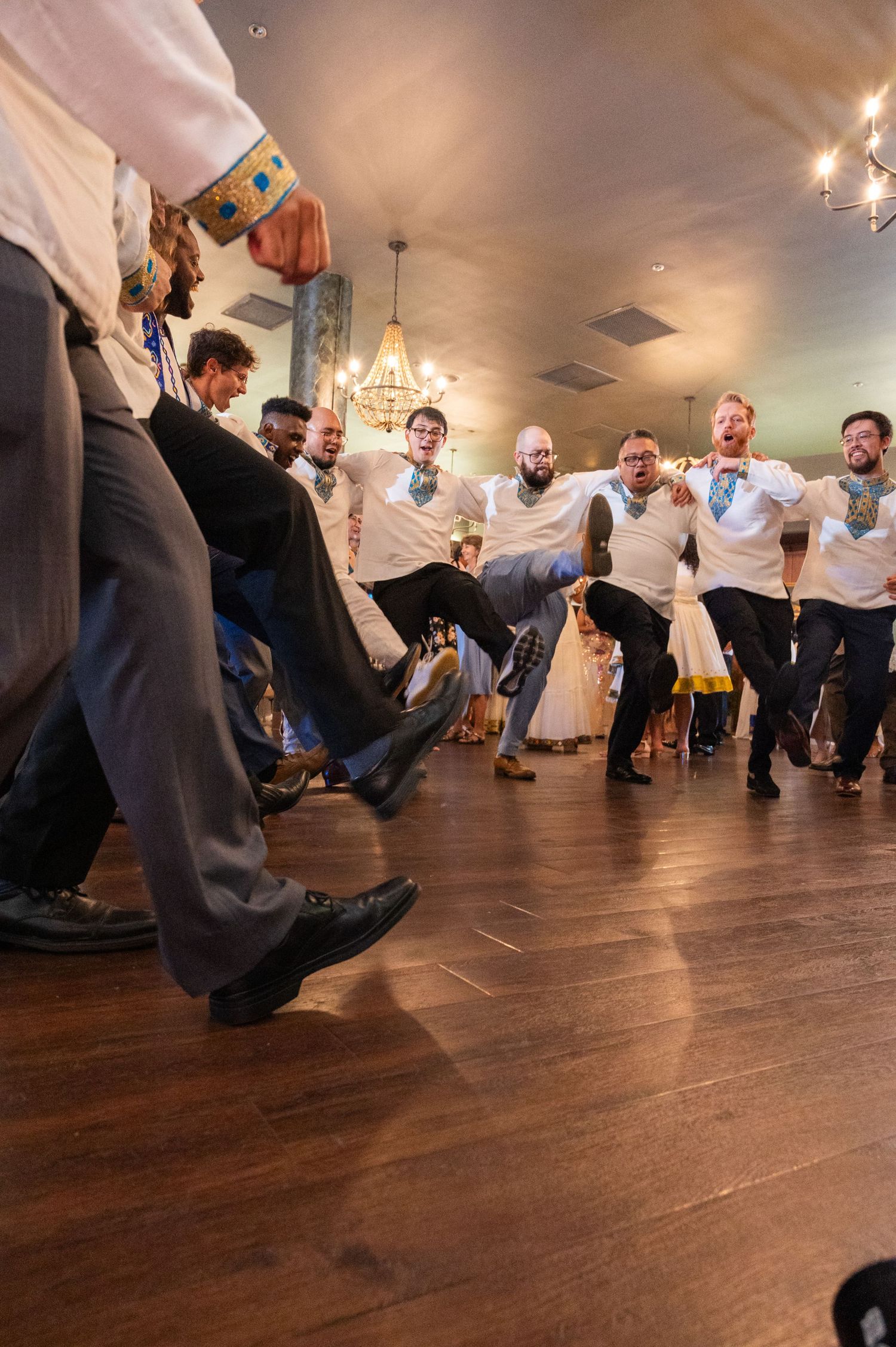 Groomsmen in white shirts perform energetic dance number on wooden dance floor at wedding reception.