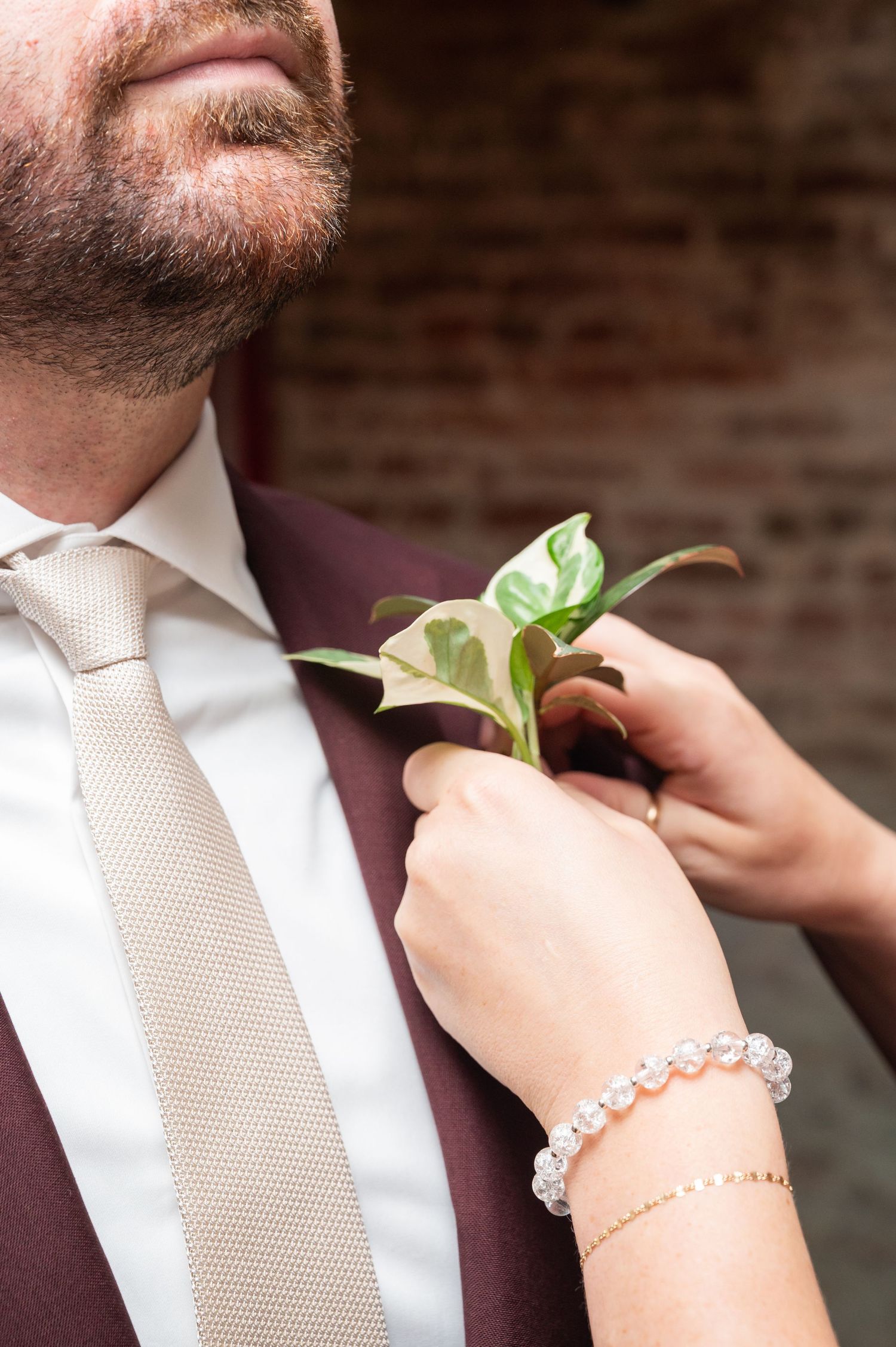 Close-up of hands pinning a green leaf boutonniere on a groom wearing burgundy suit and beige tie.