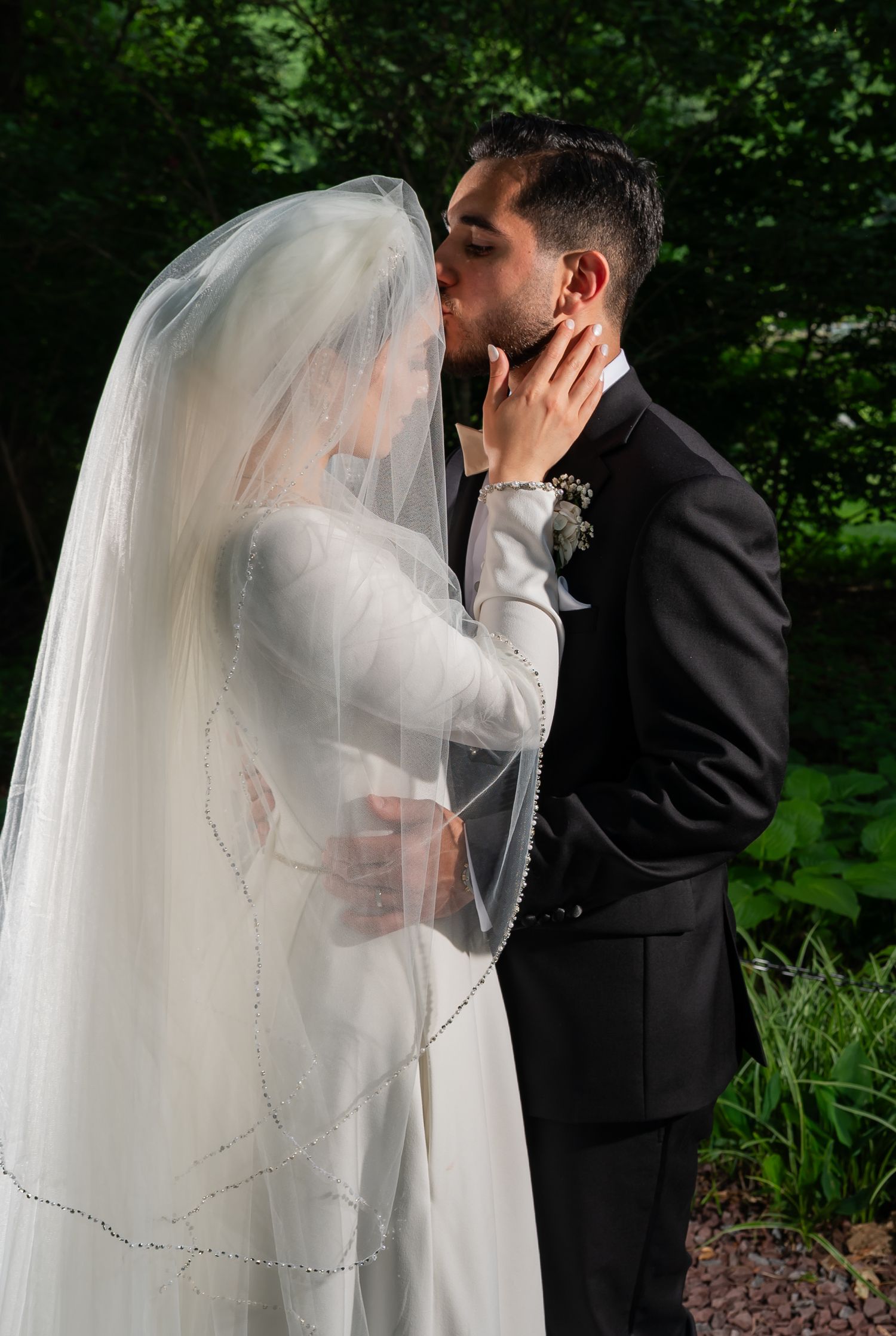 Bride and groom sharing a romantic kiss outdoors, bride wearing a beaded veil and white gown, groom in black tuxedo.