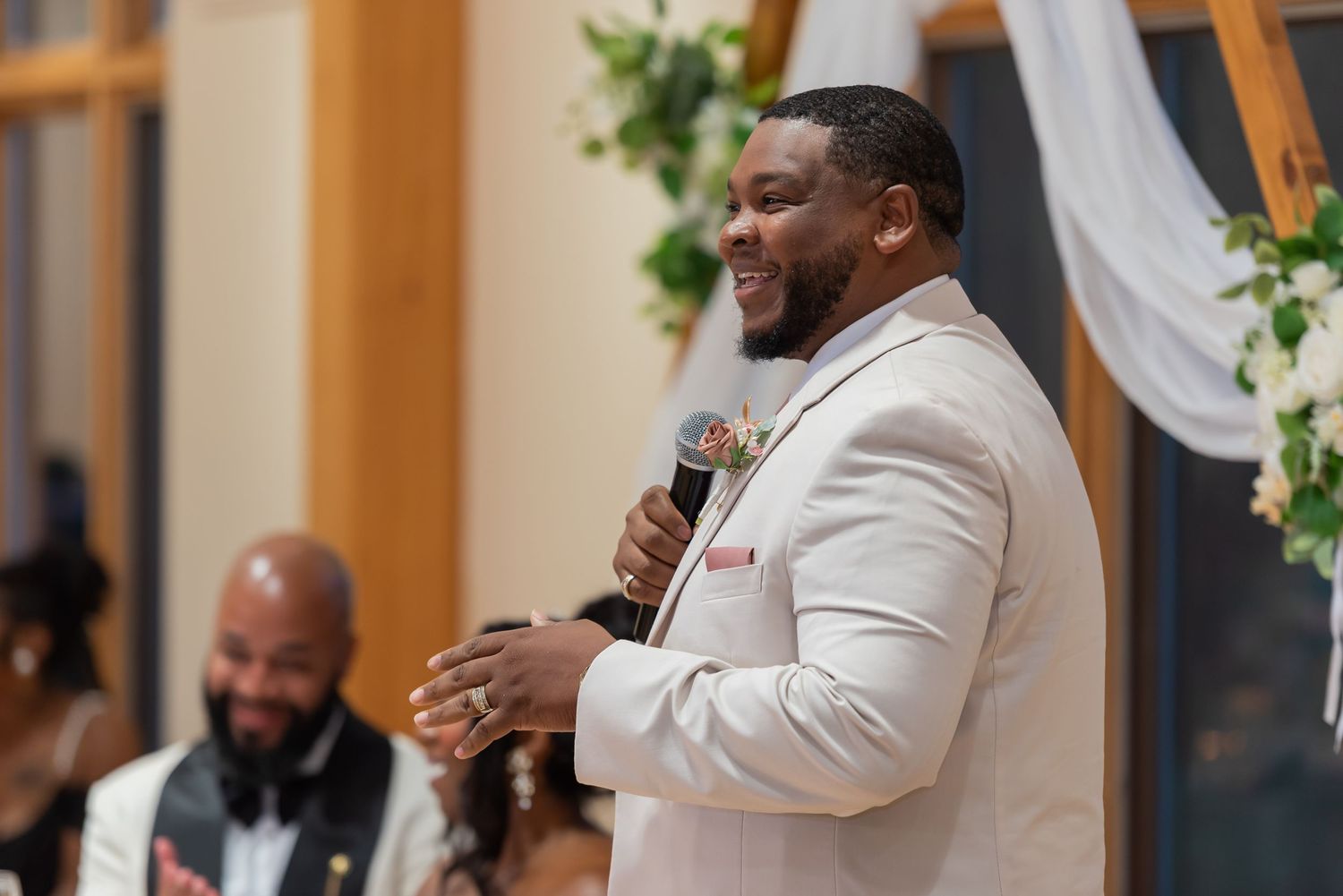 A man in a white suit smiles while speaking into a microphone at a wedding reception.