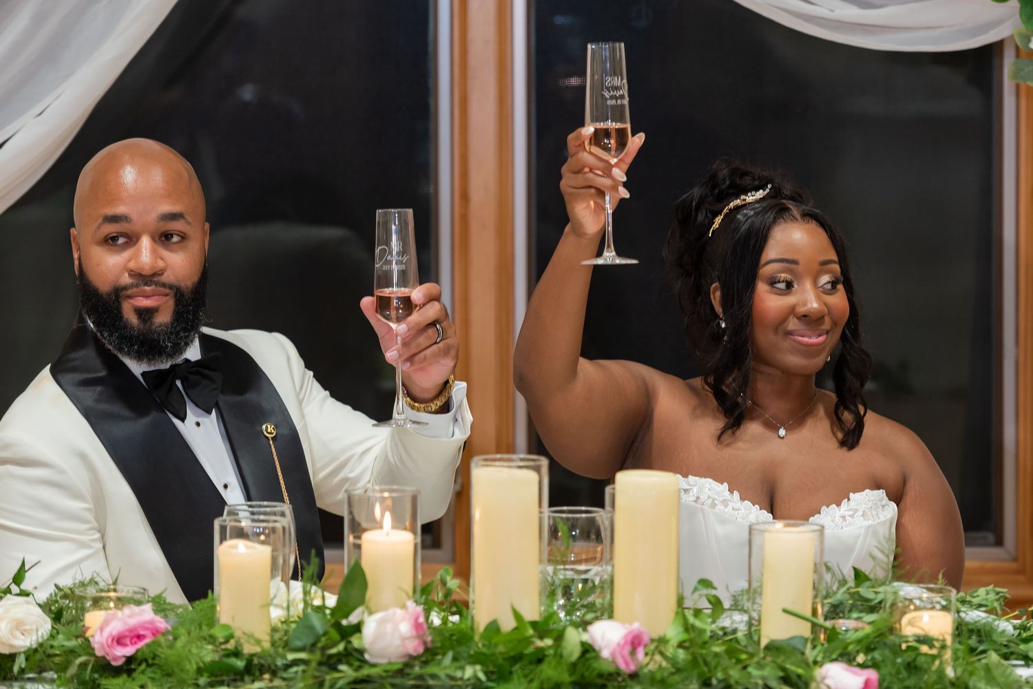 Bride and groom raising champagne glasses in a toast at their wedding reception, surrounded by candles and floral centerpieces.