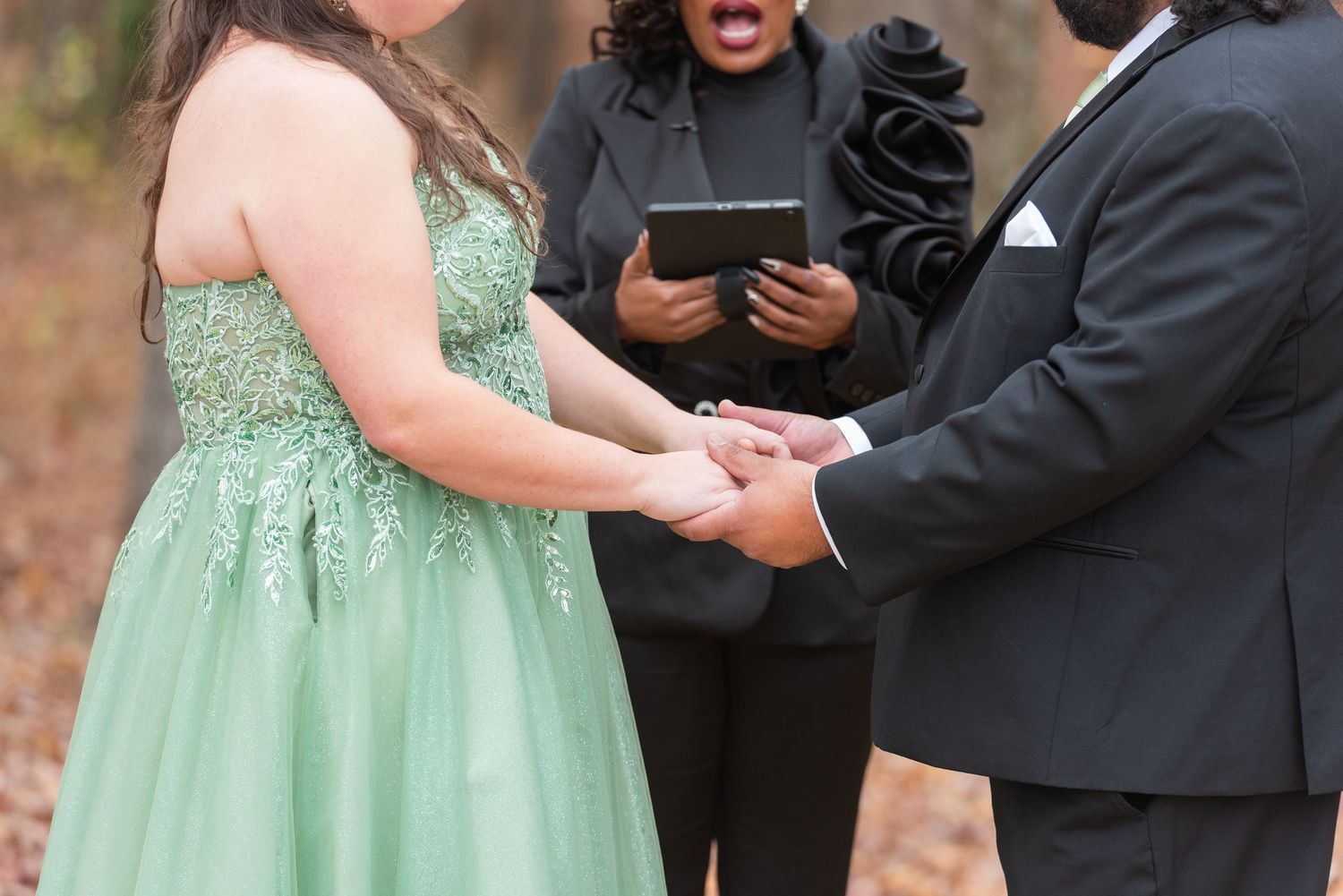 Bride in green lace gown holds hands with groom during outdoor wedding ceremony with officiant reading vows.