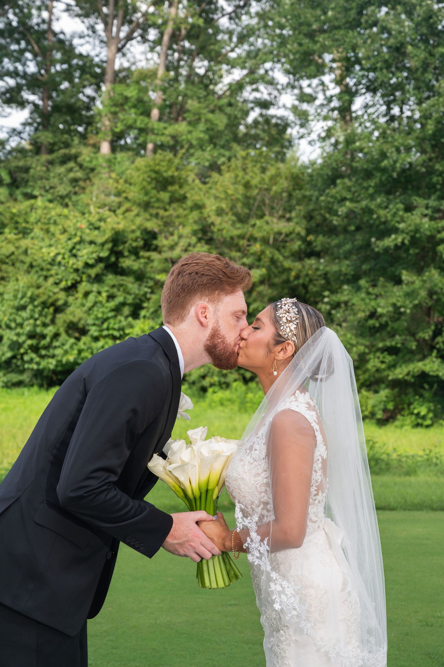 Bride and groom share a romantic kiss outdoors, bride holding white calla lily bouquet in lace gown with veil.