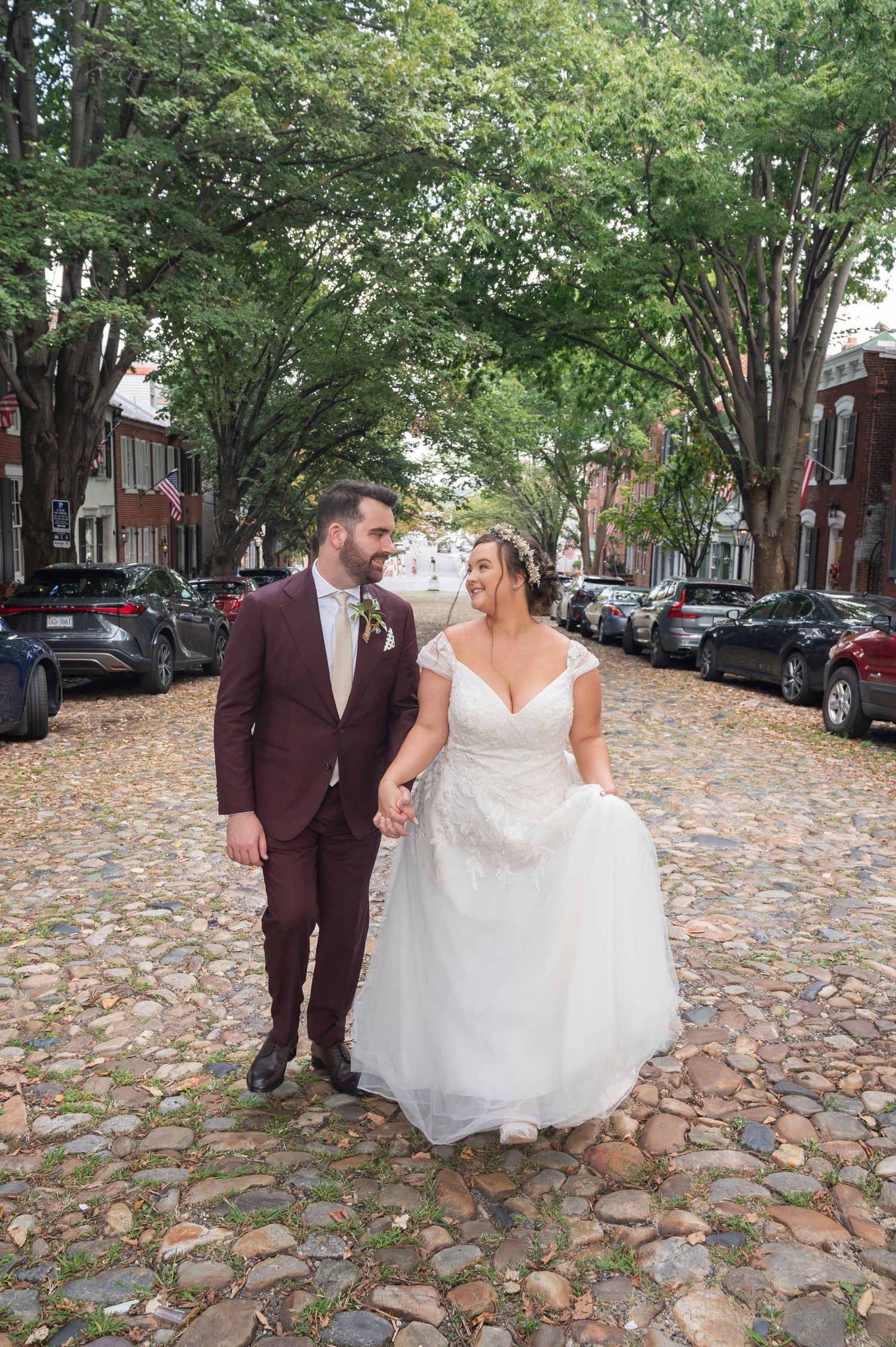 Bride and groom walking hand in hand down a charming cobblestone street lined with trees.