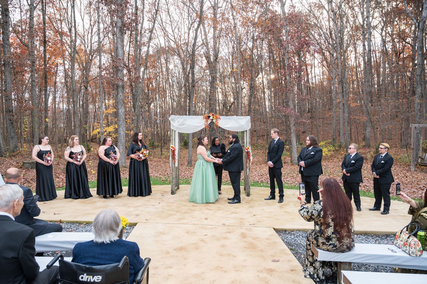 Outdoor fall wedding ceremony with bridesmaids in black gowns, couple at floral arch, groomsmen, and seated guests amid autumn trees.