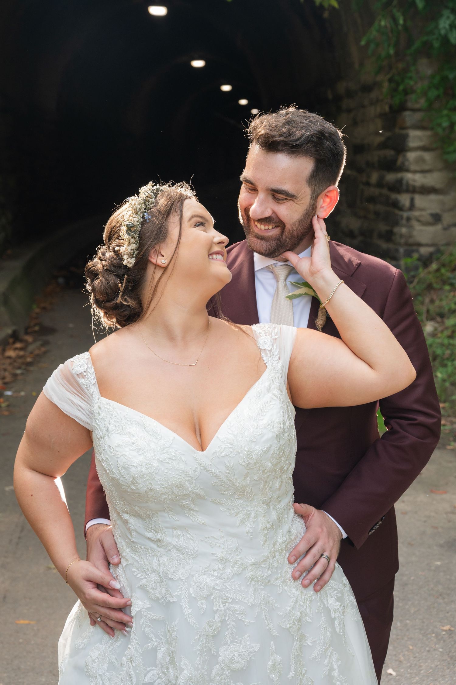 Joyful bride and groom sharing a romantic moment in a tunnel, she in lace gown, he in burgundy suit.