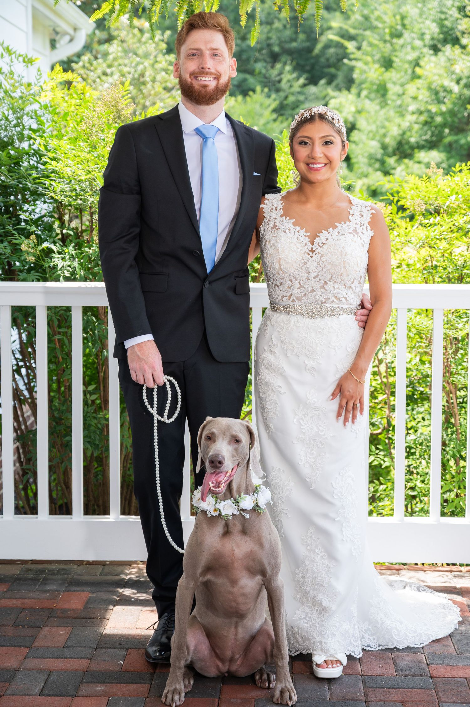 Smiling bride and groom pose with their Weimaraner dog wearing a floral collar on a white porch.
