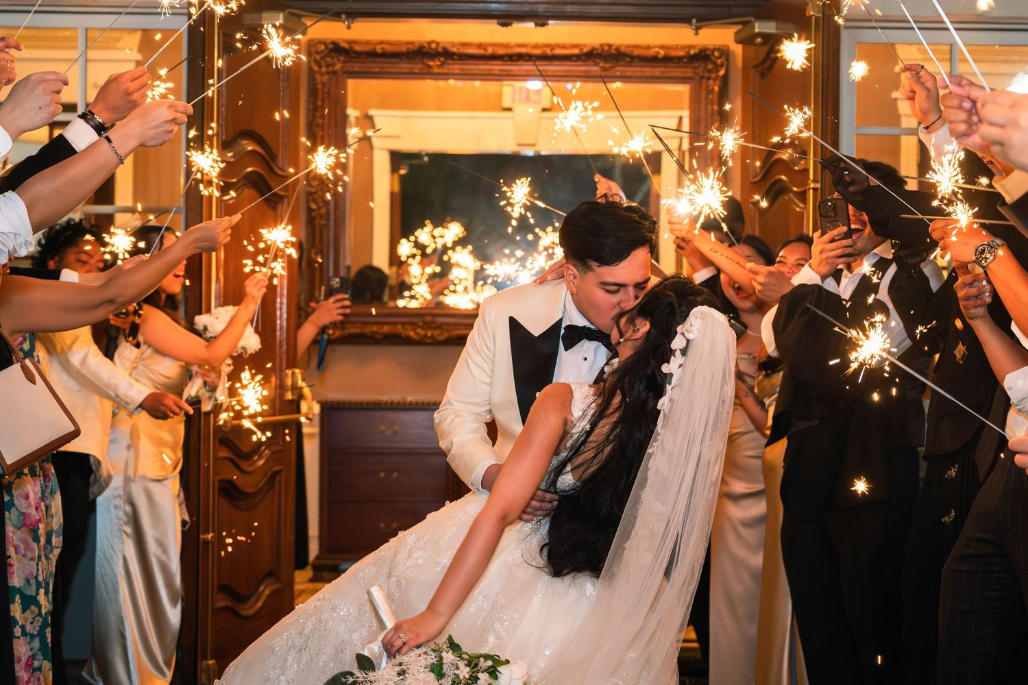Bride and groom share a romantic dip kiss surrounded by guests holding sparklers during wedding sendoff.